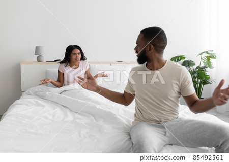 Unhappy angry excited millennial african american wife and husband swears and yells on bed in white bedroom Unhappy angry excited millennial african american wife and husband swears and yells on bed in white bedroom 85492561