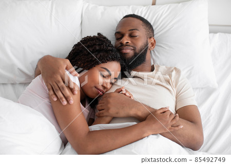 Happy young black husband hugs his wife and lie on bed on soft pillow under white blanket in bedroom Happy young black husband hugs his wife and lie on bed on soft pillow under white blanket in bedroom 85492749