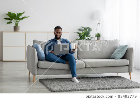 Smiling attractive millennial african american man sits on sofa drinking coffee from cup and works on pc alone 85492779