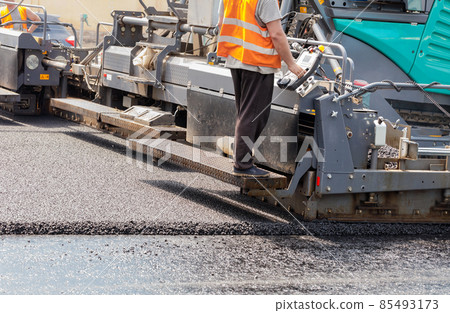 A road worker operates a tracked asphalt paver while paving fresh asphalt on the road on a summer day. 85493173