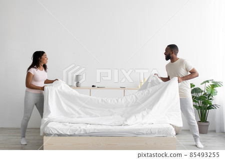 Cheerful young black woman and guy make bed and hold blanket in bedroom interior Cheerful young black woman and guy make bed and hold blanket in bedroom interior 85493255
