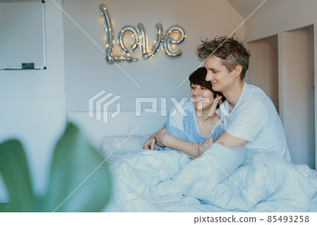 Loving couple in pajamas having fun in bed in the bedroom with word Love on the wall background. They are embracing and laughing. Happy family mornings at home. Valentine's day. Selective focus. 85493258