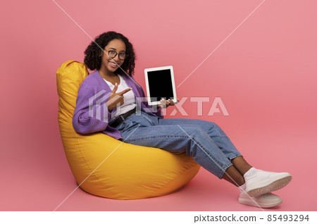 Young African American woman showing tablet pc with empty screen, sitting in bean bag chair on pink background, mockup Young African American woman showing tablet pc with empty screen, sitting in bean bag chair on pink background, mockup 85493294