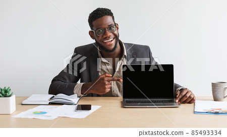 Happy young black businessman sitting at desk, pointing at laptop computer with blank screen at home office, mockup 85493334