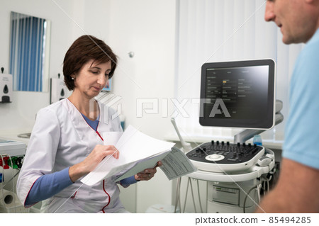 Female doctor consults male patient holding test results in hands in office of clinic. Healtcare and assistance concept. Medical exam. Woman therapist talks to client during consultation at hospital 85494285