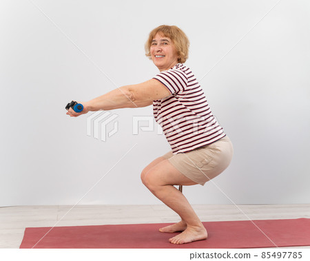 Elderly woman doing squats on a white background. The old lady is doing exercises for her health 85497785