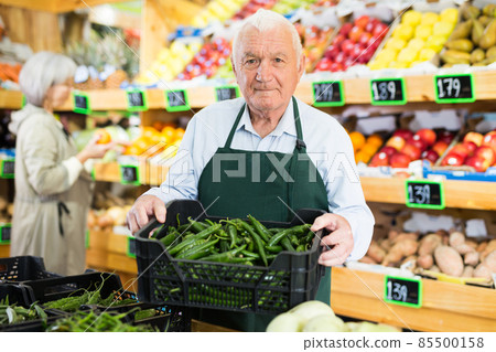 Elderly supermarket employee rearranges box of green chili peppers 85500158