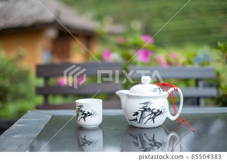 teapot and hot tea to cup on wood table against tea garden view background in the morning, Ban Rak Thai village, Mae Hong Son province, Thailand teapot and hot tea to cup on wood table against tea garden view background in the morning, Ban Rak Thai village, Mae Hong Son province, Thailand 85504383