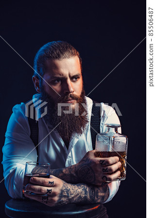 Dramatic portrait of bearded man who holds whiskey in his hands, dressed in white shirt. Studio shot 85506487