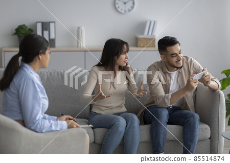 Smartphone addiction. Arab wife complaining to family counselor about her careless spouse busy with cellphone game Smartphone addiction. Arab wife complaining to family counselor about her careless spouse busy with cellphone game 85514874