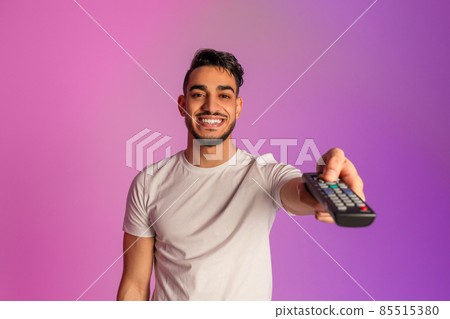 Positive young Arab guy watching TV, switching channels with remote control in neon light Positive young Arab guy watching TV, switching channels with remote control in neon light 85515380