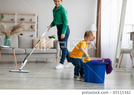Little helper. Adorable toddler boy helping mom cleaning at home, kid getting rag out of bucket while mom mopping floor 85515382