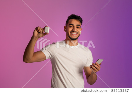 Smiling Arab guy with smartphone advertising wireless earphopnes, listening to music via new mobile app in neon light Smiling Arab guy with smartphone advertising wireless earphopnes, listening to music via new mobile app in neon light 85515387