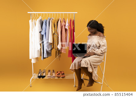 Beautiful young black woman sitting near clothes rack with laptop, offering mockup for web store design on screen Beautiful young black woman sitting near clothes rack with laptop, offering mockup for web store design on screen 85515404