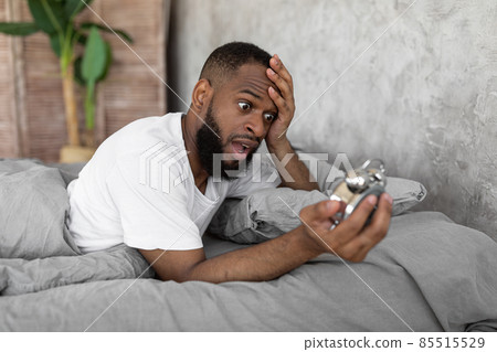 Shocked black man looking at clock in bed Shocked black man looking at clock in bed 85515529