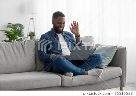 Happy attractive young african american male with beard sits on couch, greets, waves his hand to pc webcam Happy attractive young african american male with beard sits on couch, greets, waves his hand to pc webcam 85515588