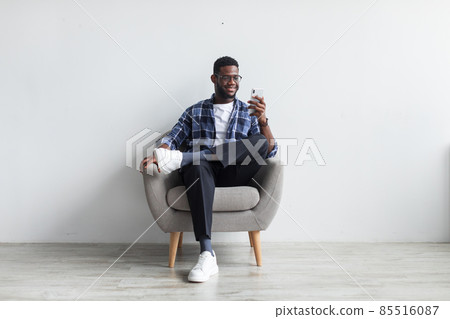 Happy black guy sitting in armchair with smartphone, texting to friend, sending message, browsing web against white wall Happy black guy sitting in armchair with smartphone, texting to friend, sending message, browsing web against white wall 85516087