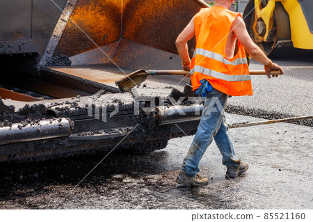 A worker scrapes fresh asphalt off a paver bucket with a shovel on a sunny day. 85521160