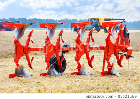 A metal multi-row plow of red color on a blurred background of wheat harvesting with large combines. A metal multi-row plow of red color on a blurred background of wheat harvesting with large combines. 85521169