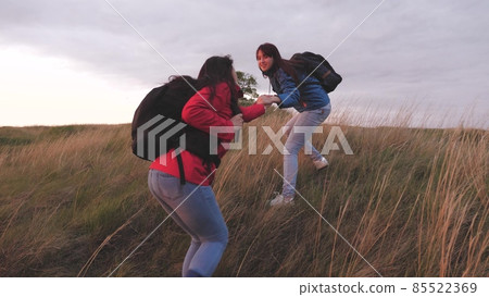 Girls with backpacks climb high up the mountain helping each other on a hiking trip. Women spend the weekend traveling in nature overcoming obstacles. Teamwork. Friends are looking for adventure 85522369