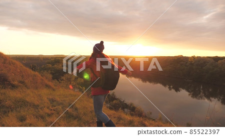A young girl with her arms outstretched feels freedom in the sunset. Woman adventurer on the background of the sky. Research work with a backpack. Weekend hike. Tourist on the ground. Life joy in 85522397