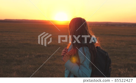 A girl traveler walks with a backpack on her back and smiles at the sunset in the sky. A teenager on a camping trip on vacation. Childhood in search of adventure from morning to dawn. Love for the 85522427