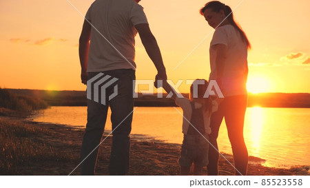 A small child holds the hand of mom and dad at the sunset sky. Childhood with parents at dawn. Happy family life. Mother, father and kid in the glare of the sun. Travel along the beach, near the coast A small child holds the hand of mom and dad at the sunset sky. Childhood with parents at dawn. Happy family life. Mother, father and kid in the glare of the sun. Travel along the beach, near the coast 85523558