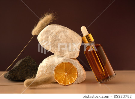Transparent glass cosmetic dropper standing near stones and dry flowers on dark red and trendy beige background. Beauty composition. Concept of natural and original skincare products presentation.  85523792