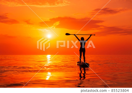 Silhouette of woman on stand up paddle board at quiet sea with sunset or sunrise. Woman posing on SUP board and bright sunset Silhouette of woman on stand up paddle board at quiet sea with sunset or sunrise. Woman posing on SUP board and bright sunset 85523804