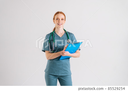 Portrait of cheerful young adult woman nurse in medical uniform writing prescription on clipboard, fills out medical history, looking at camera. 85529254