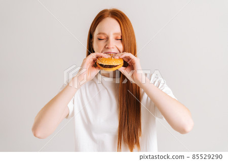 Close-up portrait of pretty young woman with closed eyes enjoying bite of appetizing delicious hamburger on white isolated background in studio. 85529290