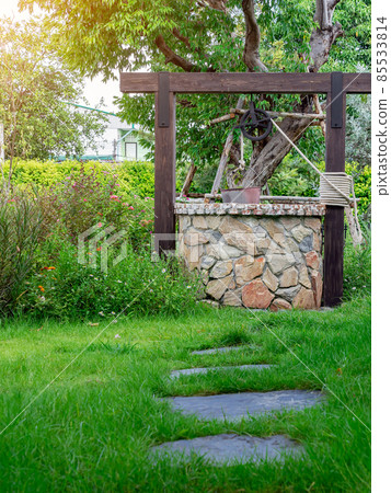 Beautiful artesian well made by stones and wheel pulley with metal bucket and rope in peaceful garden atmosphere. Retro stone water well in rural area. Garden decoration with antique items. 85533814