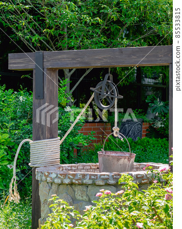 Beautiful artesian well made by stones and wheel pulley with metal bucket and rope in peaceful garden atmosphere. Retro stone water well in rural area. Garden decoration with antique items. 85533815