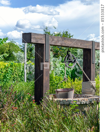 Beautiful artesian well made by stones and wheel pulley with metal bucket and rope in peaceful garden atmosphere. Retro stone water well in rural area. Garden decoration with antique items. 85533817