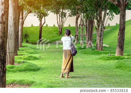 Asian female walking and use smartphone to take a selfie on pathway of park with tall green trees. A woman uses cell phone to take pictures while relaxing in the park. Happy people in green nature. 85533818