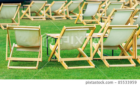 Many empty white deck chairs with tables in lawn is surrounded by shady green grass. Comfortable on outdoor patio chairs in garden. Lawn chairs in the park. Sunbeds in the garden. Selective focus. Many empty white deck chairs with tables in lawn is surrounded by shady green grass. Comfortable on outdoor patio chairs in garden. Lawn chairs in the park. Sunbeds in the garden. Selective focus. 85533959