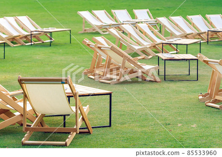 Many empty white deck chairs with tables in lawn is surrounded by shady green grass. Comfortable on outdoor patio chairs in garden.  Lawn chairs in the park. Sunbeds in the garden. Selective focus. 85533960