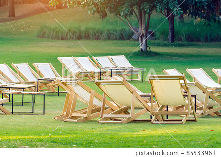 Many empty white deck chairs with tables in lawn is surrounded by shady green grass. Comfortable on outdoor patio chairs in garden.  Lawn chairs in the park. Sunbeds in the garden. Selective focus. 85533961