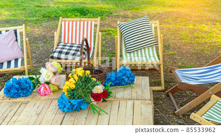 Beautiful artificial bouquets of various colors placed on wooden table among deck chairs in garden. Multi-colored artificial flowers adorn the wooden table for beautiful and relaxing. Selective focus. 85533962
