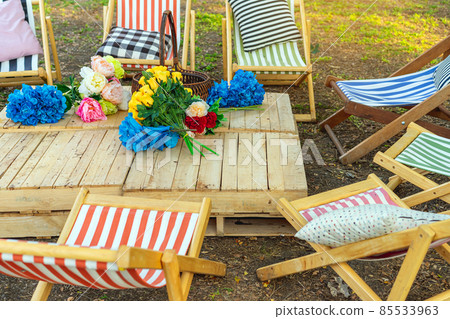 Beautiful artificial bouquets of various colors placed on wooden table among deck chairs in garden. Multi-colored artificial flowers adorn the wooden table for beautiful and relaxing. Selective focus. Beautiful artificial bouquets of various colors placed on wooden table among deck chairs in garden. Multi-colored artificial flowers adorn the wooden table for beautiful and relaxing. Selective focus. 85533963