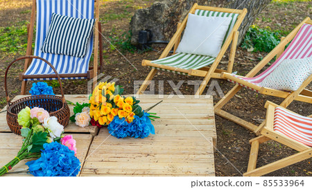 Beautiful artificial bouquets of various colors placed on wooden table among deck chairs in garden. Multi-colored artificial flowers adorn the wooden table for beautiful and relaxing. Selective focus. 85533964