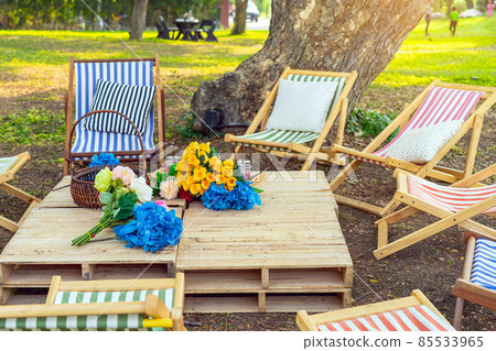 Beautiful artificial bouquets of various colors placed on wooden table among deck chairs in garden. Multi-colored artificial flowers adorn the wooden table for beautiful and relaxing. Selective focus. 85533965