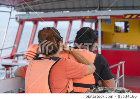 Back view of lover passengers in life vest relaxing on balcony enjoying view from boat of Samui island in sea.Happy casual couple tourists outside on tropical holiday destination.Cruise ship vacation. Back view of lover passengers in life vest relaxing on balcony enjoying view from boat of Samui island in sea.Happy casual couple tourists outside on tropical holiday destination.Cruise ship vacation. 85534160