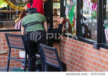 Asian women decorate beautiful stickers on window glass in front of cafe to welcome Christmas and New Year. Prepare to decorate the storefront to be beautiful during the important holidays of year. Asian women decorate beautiful stickers on window glass in front of cafe to welcome Christmas and New Year. Prepare to decorate the storefront to be beautiful during the important holidays of year. 85534382