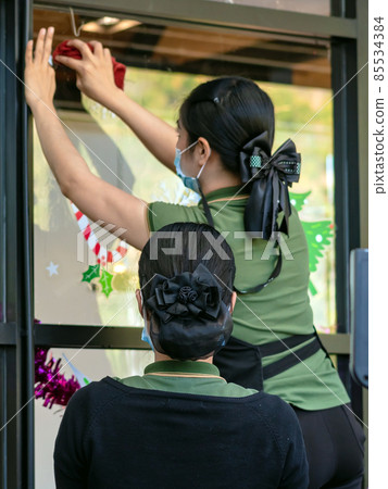 Asian women decorate beautiful stickers on window glass in front of cafe to welcome Christmas and New Year. Prepare to decorate the storefront to be beautiful during the important holidays of year. Asian women decorate beautiful stickers on window glass in front of cafe to welcome Christmas and New Year. Prepare to decorate the storefront to be beautiful during the important holidays of year. 85534384