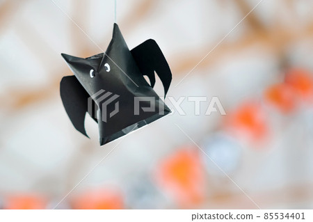 Origami bat made of black paper hanging on a rope for Halloween decorations. Dark paper ghost Halloween party concept origami paper bat. The figure of black paper bat flying over room. Space for text. 85534401