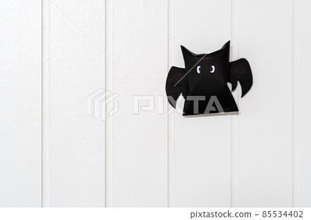 Origami bat made of black paper, isolated on white wooden wall background. Dark paper ghost Halloween party concept origami paper bat. The figure of black paper bat flying over white background. 85534402