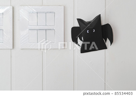 Origami bat made of black paper near light switches on white wall background. Dark paper ghost Halloween party concept origami paper bat. The figure of black paper bat flying over white background. Origami bat made of black paper near light switches on white wall background. Dark paper ghost Halloween party concept origami paper bat. The figure of black paper bat flying over white background. 85534403