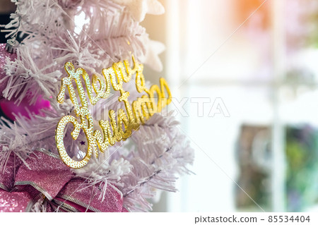 Golden shiny Merry Christmas handwriting logo on white paper tree for christmas time in coffee shop. Christmas tree decorations with color lights and a merry christmas logo in cafe. Selective focus Golden shiny Merry Christmas handwriting logo on white paper tree for christmas time in coffee shop. Christmas tree decorations with color lights and a merry christmas logo in cafe. Selective focus 85534404