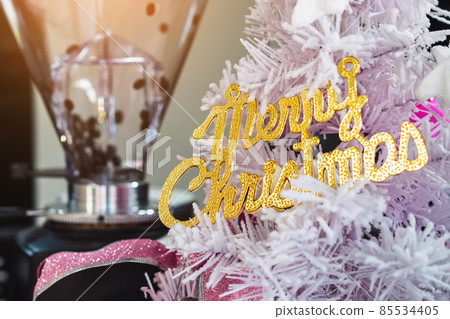 Golden shiny Merry Christmas handwriting logo on white paper tree for christmas time in coffee shop. Christmas tree decorations with color lights and a merry christmas logo in cafe. Selective focus Golden shiny Merry Christmas handwriting logo on white paper tree for christmas time in coffee shop. Christmas tree decorations with color lights and a merry christmas logo in cafe. Selective focus 85534405